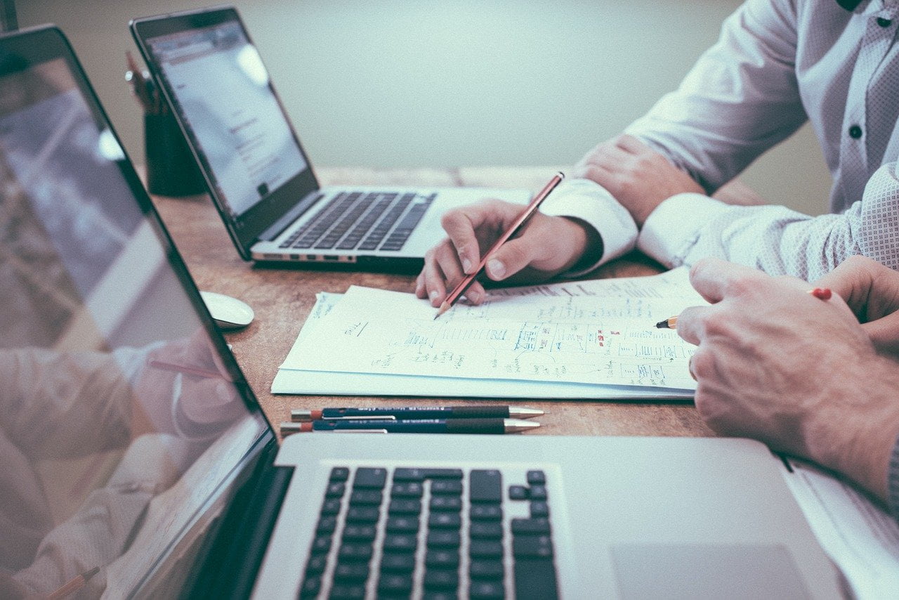 Two business men sit at a desk in front of two laptops discussing a software requirements specification