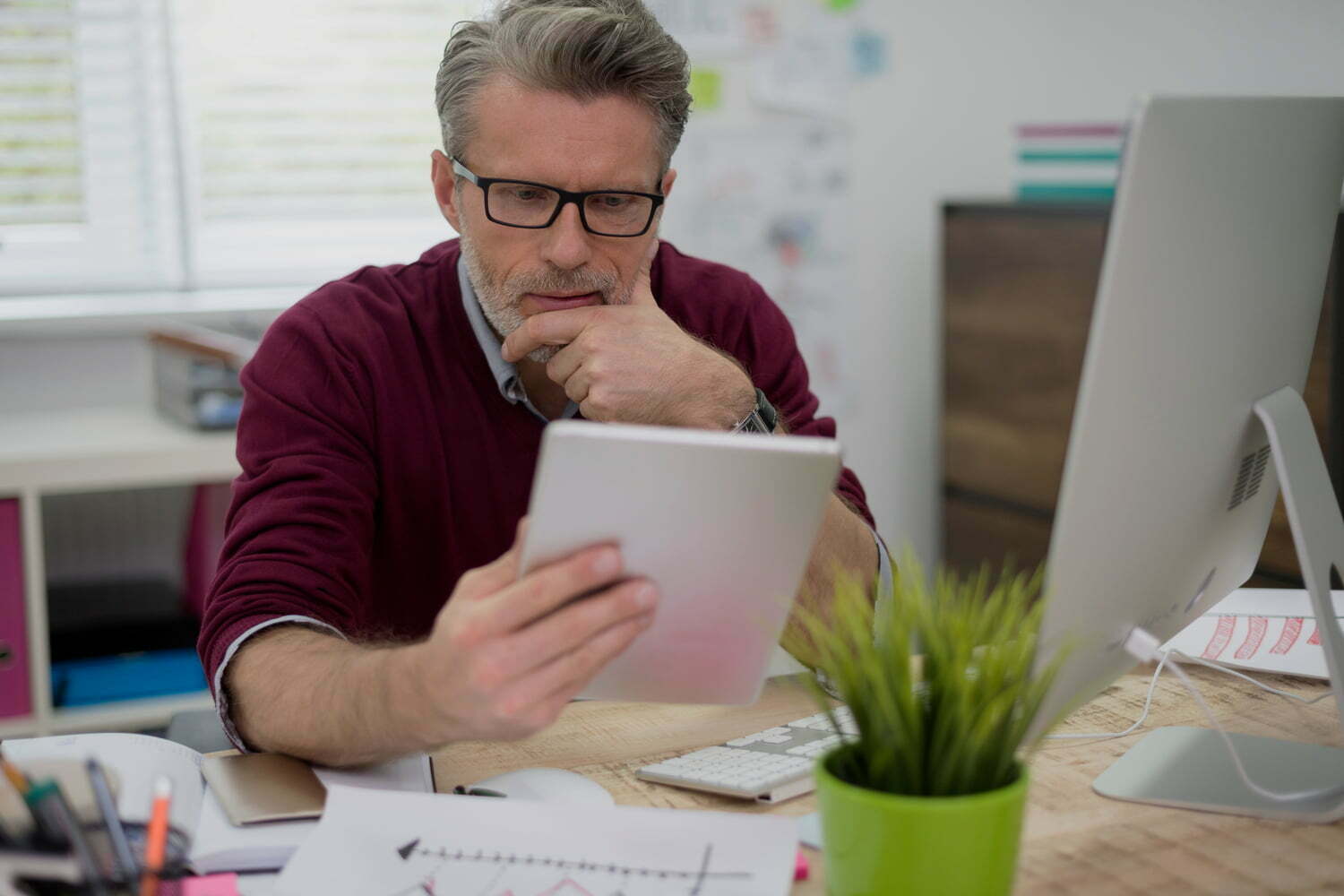 Businessman at desk with desktop computer holds tablet while thinking