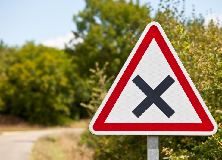 Photo of a road and sign with foliage behind it. The sign is a triangle with red outline and a large black cross.
