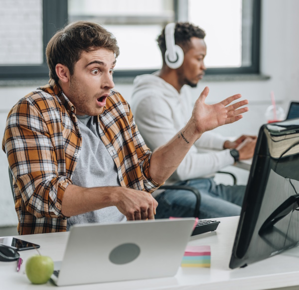 Photo of a man shocked by the computer software he is using, sat in front of a monitor and laptop.