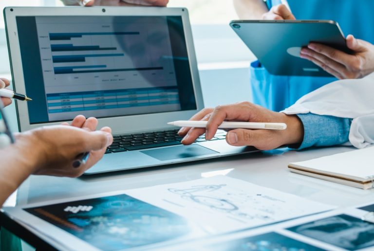 Photo of doctors hands and pens, laptop computer with healthcare software solutions on the screen.